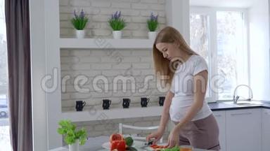 <strong>怀孕期</strong>间的营养，美丽的女孩切蔬菜和吃甜椒抱着她的大肚子在厨房